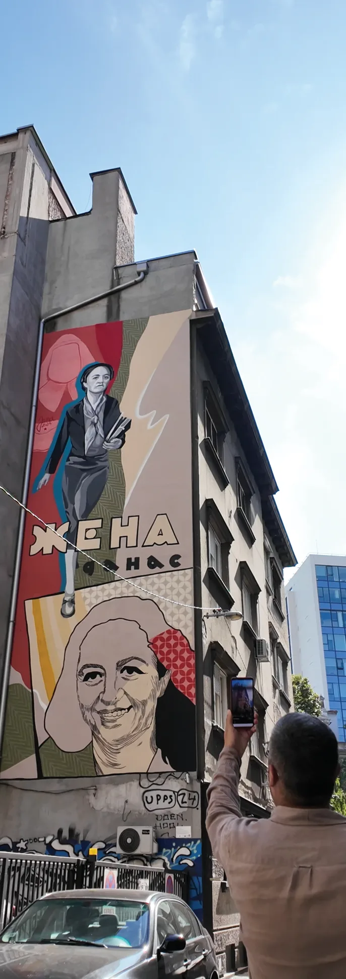 A person pointing a phone toward a tall mural on a building.